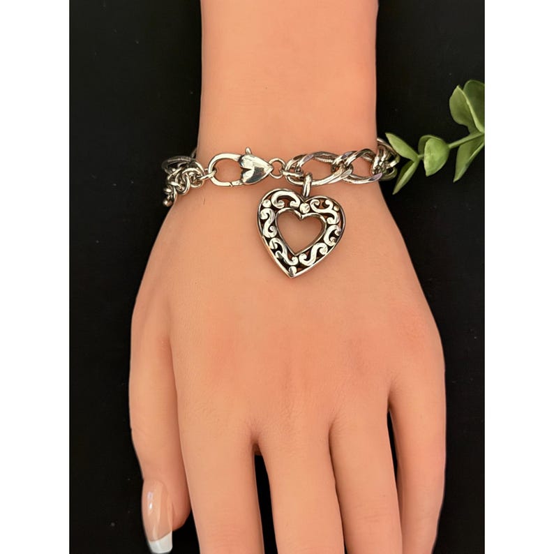Op de afbeelding: Zilverkleurige armband met een hartvormige bedel. De bedel heeft een open ontwerp met ingewikkelde krullen. De armband wordt getoond op een mannequinhand, die de stijl van de sieraden benadrukt. De ketting is een curb link stijl.