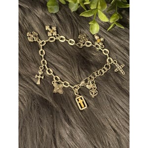 May include: Silver-toned charm bracelet featuring various cross-shaped charms and other decorative elements. The bracelet includes charms with intricate designs and a mix of textures, creating a detailed and eye-catching accessory. The bracelet is displayed on a dark, textured surface.