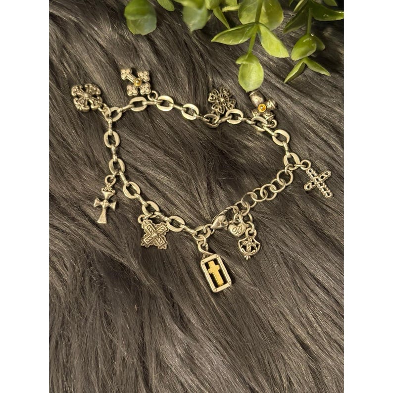 May include: Silver-toned charm bracelet featuring various cross-shaped charms and other decorative elements. The bracelet includes charms with intricate designs and a mix of textures, creating a detailed and eye-catching accessory. The bracelet is displayed on a dark, textured surface.