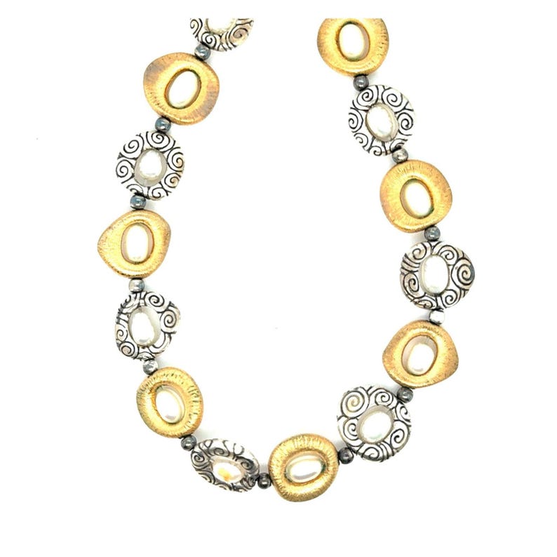 Puede incluir: Un collar con eslabones ovalados dorados y circulares plateados alternados. Los eslabones dorados tienen una abertura ovalada central, mientras que los eslabones plateados tienen un dise&ntilde;o en espiral. Peque&ntilde;as cuentas oscuras conectan los eslabones.