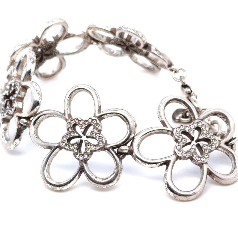 Pode incluir: Pulseira prateada com design floral. Cada flor tem cinco p&eacute;talas vazadas e um centro adornado com pequenos cristais transparentes. A pulseira &eacute; ligada com pequenos conectores circulares.