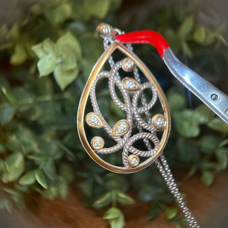 May include: A teardrop pendant necklace with a gold-tone frame and silver chain. The pendant has a textured, rope-like design with clear, round gemstones. The necklace is shown against a blurred green background.