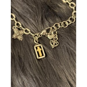 May include: Silver-toned charm bracelet with multiple religious charms. The charms include crosses, a heart-shaped charm, and a rectangular charm with a gold cross. The bracelet is displayed on a dark gray, textured surface.