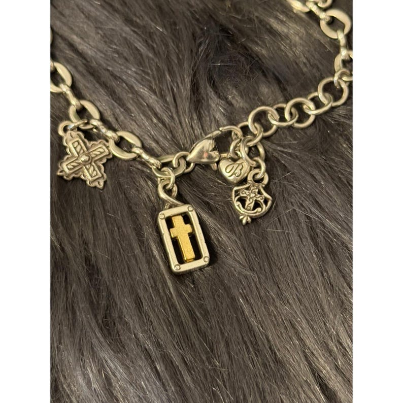 May include: Silver-toned charm bracelet with multiple religious charms. The charms include crosses, a heart-shaped charm, and a rectangular charm with a gold cross. The bracelet is displayed on a dark gray, textured surface.