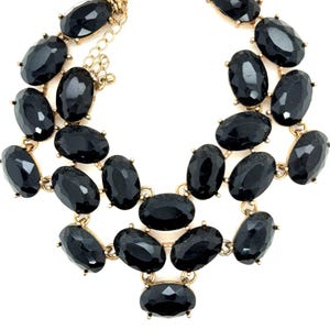 May include: A statement necklace featuring multiple faceted, oval-shaped black stones set in gold-toned metal. The stones are arranged in a layered, cascading design with a metal chain extender and a lobster claw clasp visible at the top.