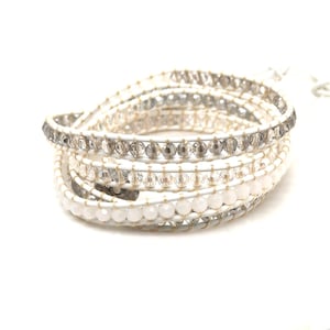 May include: A multi-strand wrap bracelet featuring white leather cord and rows of faceted beads in clear, light gray, and opaque white. The beads are secured to the leather cord with thin, light-colored thread, creating a layered, coiled appearance.