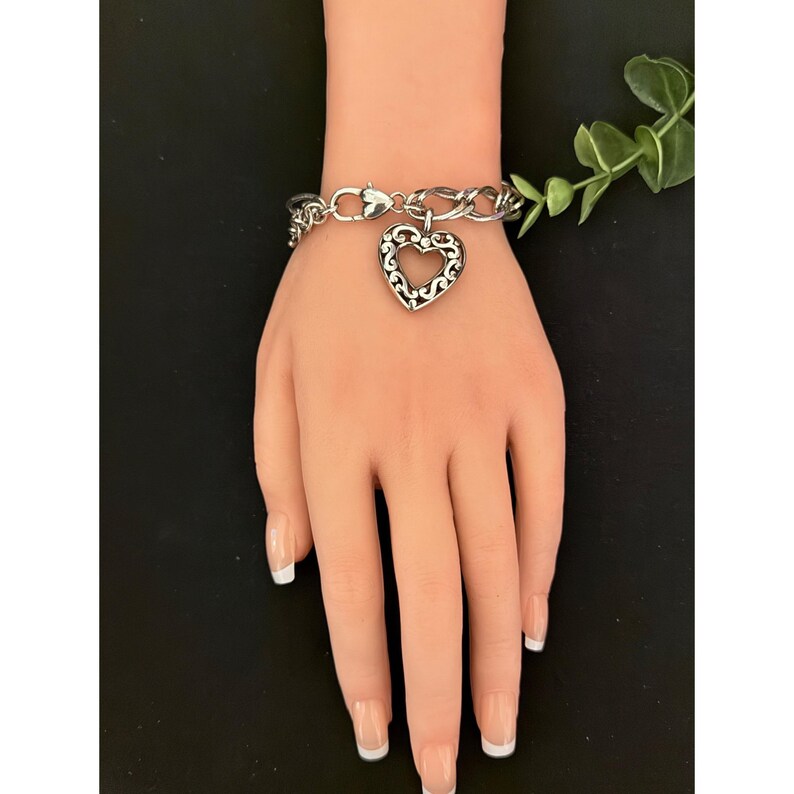 Op de afbeelding: Een zilverkleurige armband met een hartvormige bedel. De bedel heeft een filigraan ontwerp en hangt aan een schakelarmband. De armband wordt getoond op een mannequinhand met verzorgde nagels.