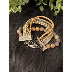 Op de afbeelding: Een armband met meerdere strengen met een geweven gouden band, een gevlochten bruinleren band en een kralenstreng. De armband heeft zilveren sluitingen met de namen "EMERSON" en "VICTORIA" gegraveerd. De kralen zijn beige en oranje.