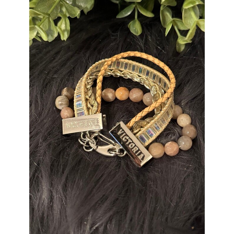 Op de afbeelding: Een armband met meerdere strengen met een geweven gouden band, een gevlochten bruinleren band en een kralenstreng. De armband heeft zilveren sluitingen met de namen "EMERSON" en "VICTORIA" gegraveerd. De kralen zijn beige en oranje.