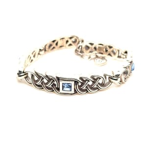 May include: Silver-toned bracelet featuring a Celtic knot design. The bracelet has a series of interwoven knots with square, light blue gemstones. The bracelet is on a white background.