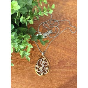 May include: A silver necklace with a gold-toned teardrop pendant. The pendant has a swirling design with small, clear gemstones. The necklace is displayed on a wooden surface with green foliage.