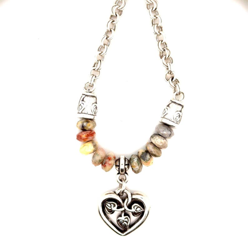 May include: A silver necklace with a heart-shaped pendant. The pendant has a leaf design. The necklace features a chain and a strand of colorful, irregularly shaped beads. The beads are in shades of red, yellow, and gray.