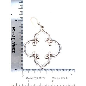 May include: A silver-coloured earring with a quatrefoil design. The earring is approximately 5 cm long. The design features curved lines and scroll-like details. The earring is made of stainless steel and made in the U.S.A.