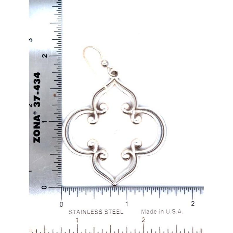 May include: A silver-coloured earring with a quatrefoil design. The earring is approximately 5 cm long. The design features curved lines and scroll-like details. The earring is made of stainless steel and made in the U.S.A.