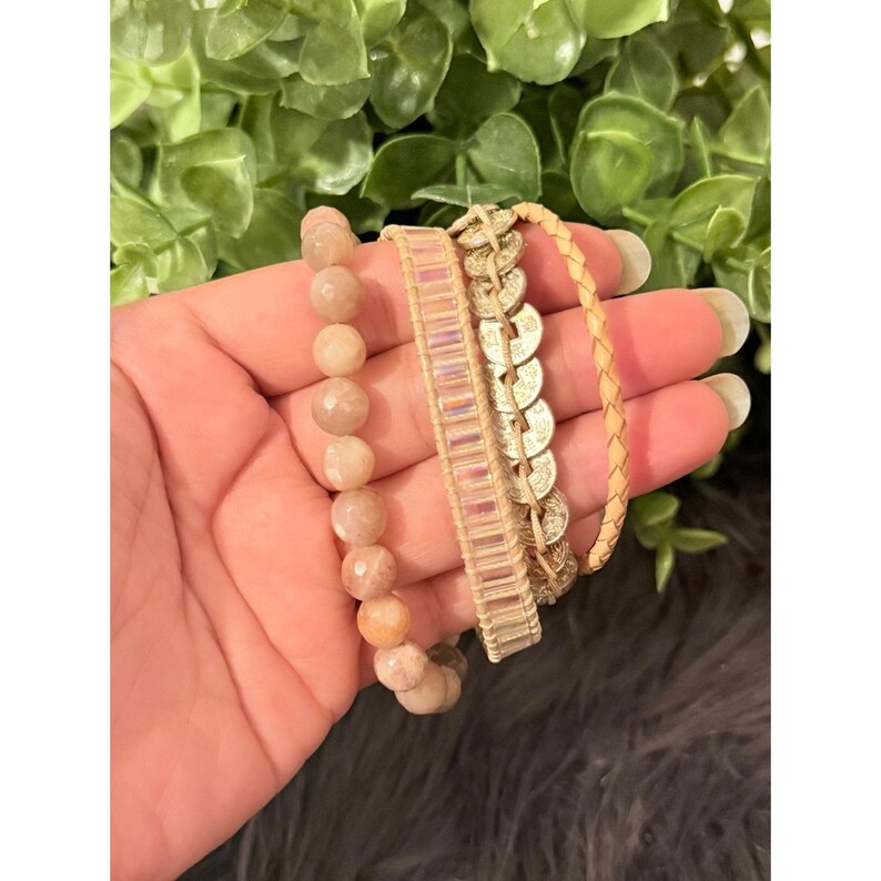 Op de afbeelding: Een collectie armbanden in verschillende stijlen. E&eacute;n armband heeft ronde, perzik-kleurige kralen. Een andere heeft een geweven ontwerp met iriserende rechthoekige kralen. De derde is een gevlochten, lichtbruine leren armband. De armbanden zijn op elkaar gestapeld.