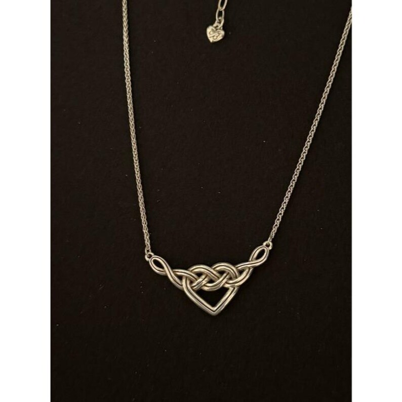 May include: Silver-toned necklace featuring a heart-shaped pendant with an interwoven knot design. The pendant is connected to a delicate chain, and the necklace has a small heart charm near the clasp. The jewelry is displayed against a black background.