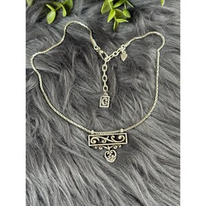 May include: Silver-toned necklace with a decorative rectangular pendant featuring a scrolled design and a heart-shaped accent. The necklace has a delicate chain and an adjustable length.