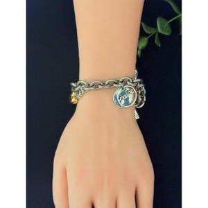 May include: Silver-toned chain bracelet with multiple charms. One charm features the word "peace" in a blue and white design. Additional charms include a gold-colored bead and a small silver charm. The bracelet is displayed on a light skin tone.