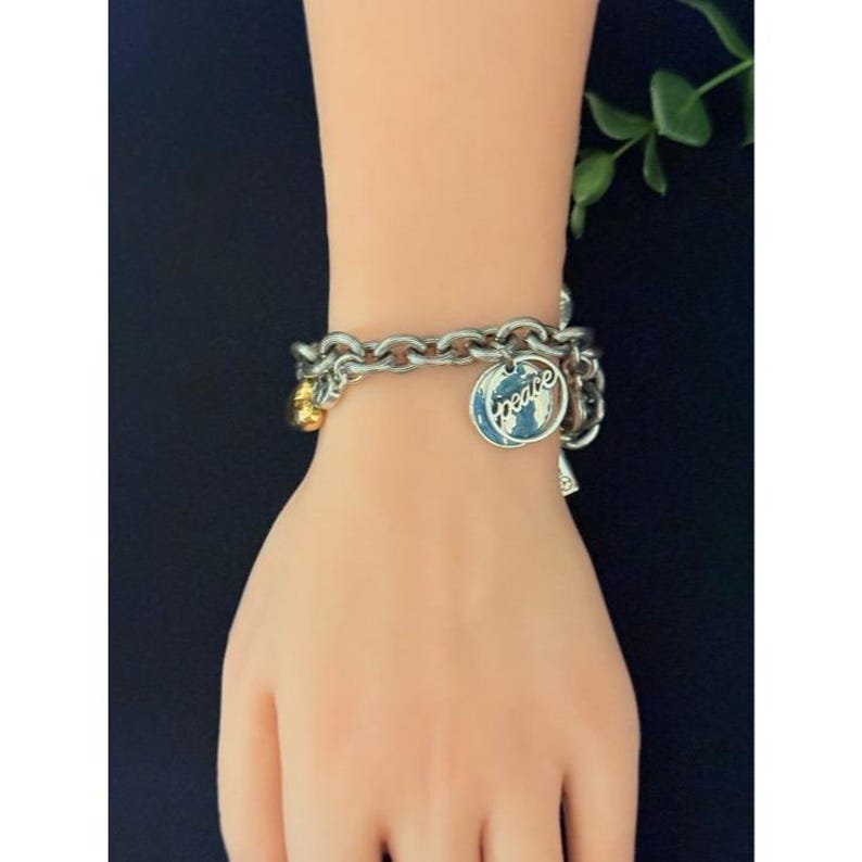 May include: Silver-toned chain bracelet with multiple charms. One charm features the word "peace" in a blue and white design. Additional charms include a gold-colored bead and a small silver charm. The bracelet is displayed on a light skin tone.
