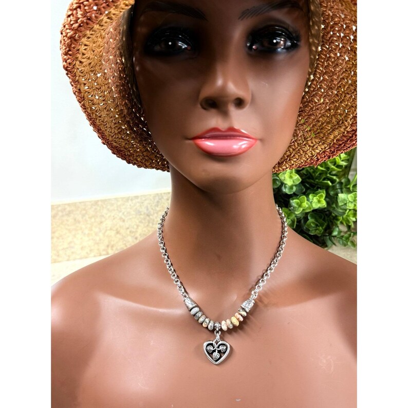 May include: Silver-toned necklace with a heart-shaped pendant. The pendant features a decorative design. The necklace is displayed on a mannequin wearing a straw hat.