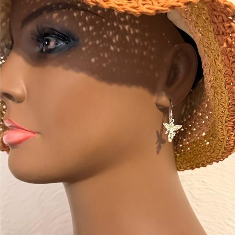 May include: A silver-colored bee-shaped earring is shown hanging from an ear. The earring is detailed with wings and a body. The background includes a straw hat and the side profile of a mannequin.