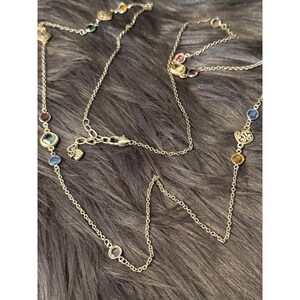 K&ouml;nnte beinhalten: Eine silberfarbene Halskette mit bunten Edelsteinen. Die Kette hat eine filigrane Kette mit Edelsteinen in Blau, Rot, Gr&uuml;n und Gelb. Die Edelsteine haben verschiedene Formen und Gr&ouml;&szlig;en. Die Kette liegt auf einer dunkelgrauen, strukturierten Oberfl&auml;che.