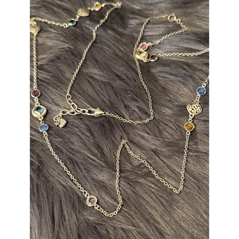 K&ouml;nnte beinhalten: Eine silberfarbene Halskette mit bunten Edelsteinen. Die Kette hat eine filigrane Kette mit Edelsteinen in Blau, Rot, Gr&uuml;n und Gelb. Die Edelsteine haben verschiedene Formen und Gr&ouml;&szlig;en. Die Kette liegt auf einer dunkelgrauen, strukturierten Oberfl&auml;che.