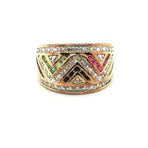 May include: A wide gold band ring featuring a geometric chevron pattern. The design is accented with rows of small white diamonds, green gemstones, red rubies, and dark blue sapphires set within the triangular sections of the metal band.