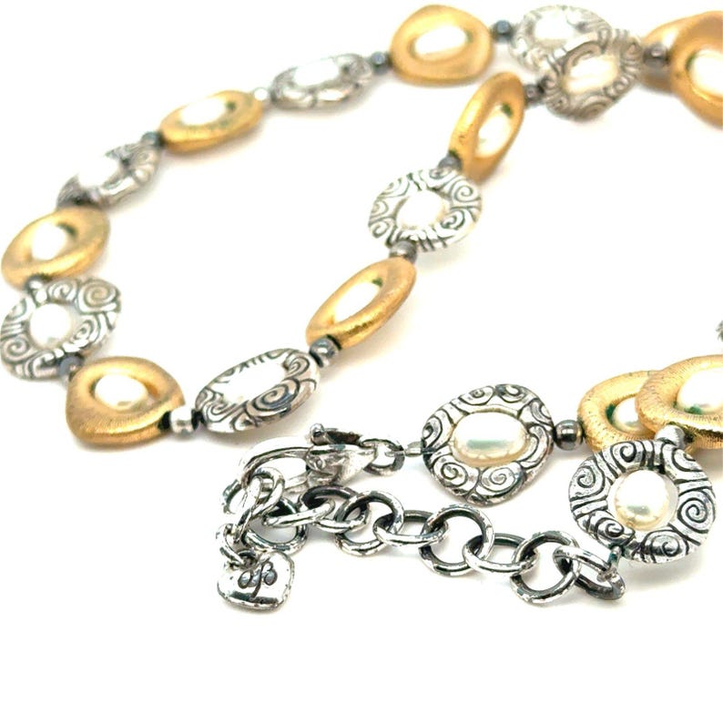 Puede incluir: Un collar plateado y dorado con una variedad de eslabones ovalados y circulares. El collar presenta una mezcla de texturas, que incluyen metal dorado liso, detalles plateados ornamentados y peque&ntilde;as perlas redondas.