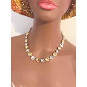 Puede incluir: Un collar dorado y plateado con un dise&ntilde;o circular. El collar presenta eslabones circulares dorados y plateados alternados, algunos con un centro similar a una perla. El collar se exhibe en un maniqu&iacute;.