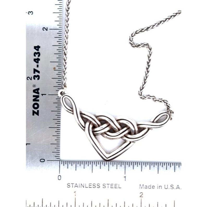 May include: A silver-toned Celtic knot pendant necklace. The pendant features an intricate interwoven design, with a heart-shaped element at the bottom. The chain is a delicate, braided style. A ruler is placed next to the necklace, showing measurements in inches.