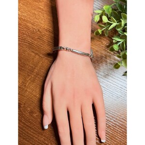 Puede incluir: Una pulsera plateada con un dise&ntilde;o decorativo se lleva en la mu&ntilde;eca. La pulsera tiene un cierre rectangular y una secci&oacute;n central texturizada. La mano est&aacute; sobre una superficie de madera, con vegetaci&oacute;n en el fondo.