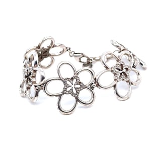 Pode incluir: Pulseira prateada com desenhos florais vazados. Cada flor tem cinco p&eacute;talas e um centro brilhante. A pulseira &eacute; conectada por uma delicada corrente e fecho, criando um acess&oacute;rio floral.