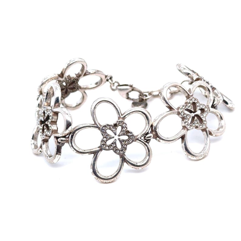 Pode incluir: Pulseira prateada com desenhos florais vazados. Cada flor tem cinco p&eacute;talas e um centro brilhante. A pulseira &eacute; conectada por uma delicada corrente e fecho, criando um acess&oacute;rio floral.