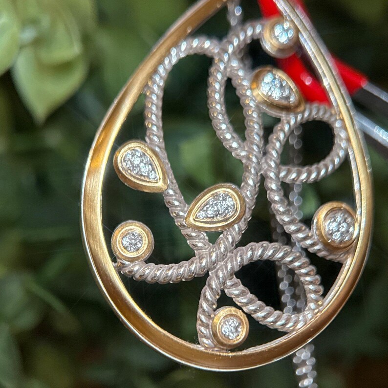 May include: A teardrop-shaped pendant with a gold border and silver interior. The pendant has a rope-like design with small, round and teardrop-shaped gold accents, each set with small, sparkling stones. The background is a soft green.