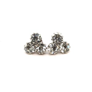 May include: A pair of silver-toned stud earrings, each featuring three clear, round, faceted crystals arranged in a triangular cluster. The earrings are set against a white background, highlighting their sparkle and design.