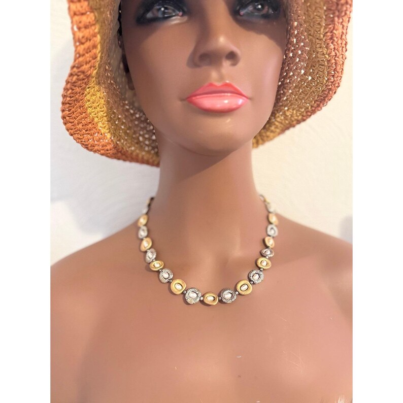 Puede incluir: Un collar dorado y plateado con una serie de colgantes circulares. Cada colgante tiene un anillo exterior dorado y un centro plateado, algunos con detalles de piedras transparentes. El collar se exhibe en un maniqu&iacute; que lleva un sombrero de paja.