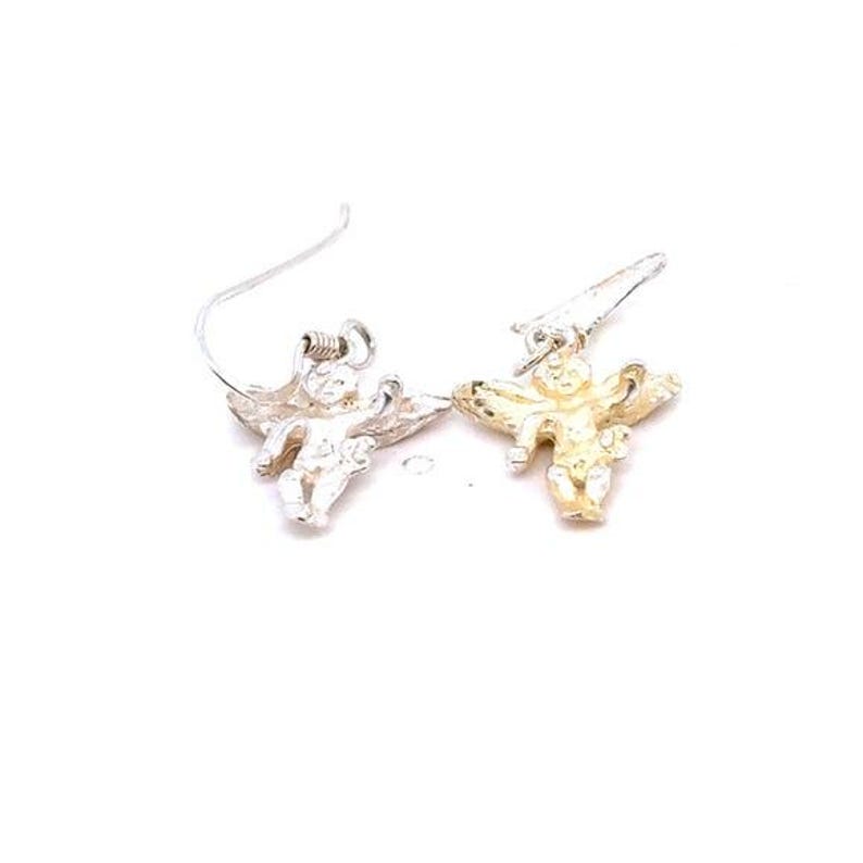 May include: A pair of angel-shaped earrings. One earring is silver-toned, the other is gold-toned. Each earring features a small cherub with outstretched wings, suspended from a curved ear wire. The earrings are set against a white background.