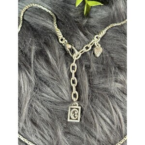May include: Silver-toned necklace with a braided chain design. The necklace features a heart-shaped charm and a rectangular pendant with a stylized letter "G". The clasp and chain links are visible.