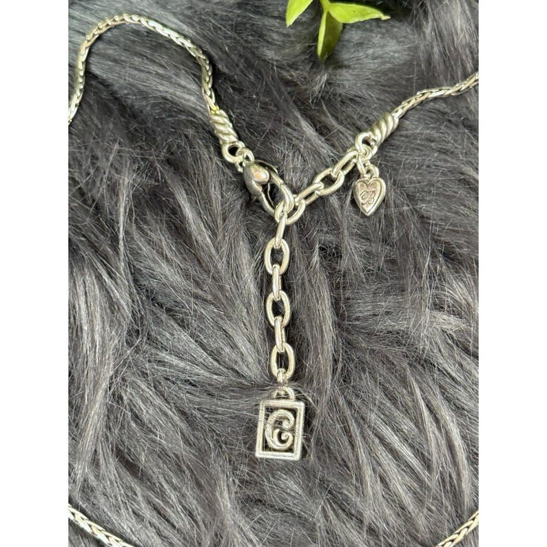 May include: Silver-toned necklace with a braided chain design. The necklace features a heart-shaped charm and a rectangular pendant with a stylized letter "G". The clasp and chain links are visible.