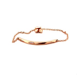 May include: A rose gold-toned bracelet featuring a curved, faceted bar centerpiece with small embedded clear stones. The bracelet includes a delicate chain and an adjustable sliding clasp mechanism with two small, pointed metal end pieces.