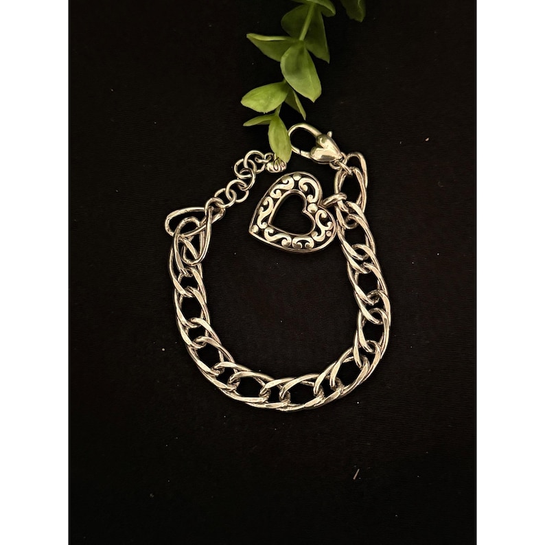Op de afbeelding: Zilverkleurige armband met een hartvormige bedel. De bedel heeft een open ontwerp. De armband heeft een schakelontwerp en een karabijnsluiting. De armband wordt getoond op een zwarte ondergrond met een groene plant.