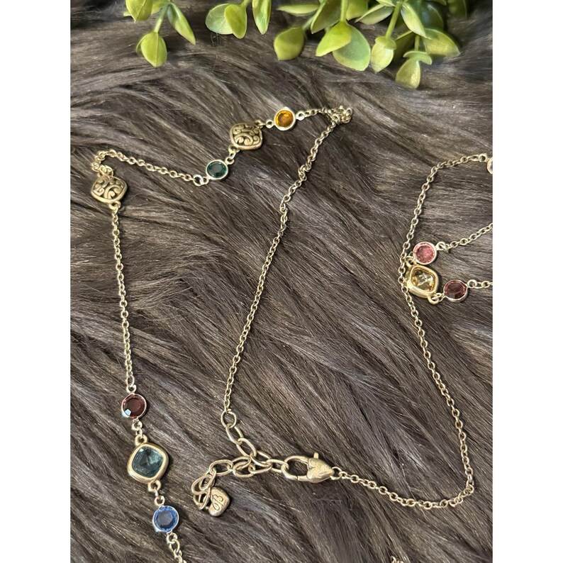 K&ouml;nnte beinhalten: Eine goldfarbene Halskette mit bunten Edelsteinen. Die Halskette hat eine zarte Kette mit verschiedenen farbigen Steinen, darunter Blau, Gr&uuml;n, Rot und Gelb. Die Edelsteine sind in goldfarbenen Fassungen gefasst. Die Halskette wird auf einer strukturierten, dunkelbraunen Oberfl&auml;che pr&auml;sentiert.