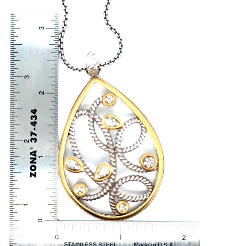 May include: A silver and gold-tone pendant necklace with a teardrop design. The pendant features a gold-tone border, silver-tone rope-like detailing, and clear gemstones. The pendant measures approximately 5 cm in length.