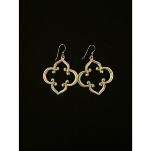 May include: A pair of gold-coloured, openwork earrings with a quatrefoil design. The earrings have a hook closure and are displayed against a black background. The earrings are a decorative accessory.