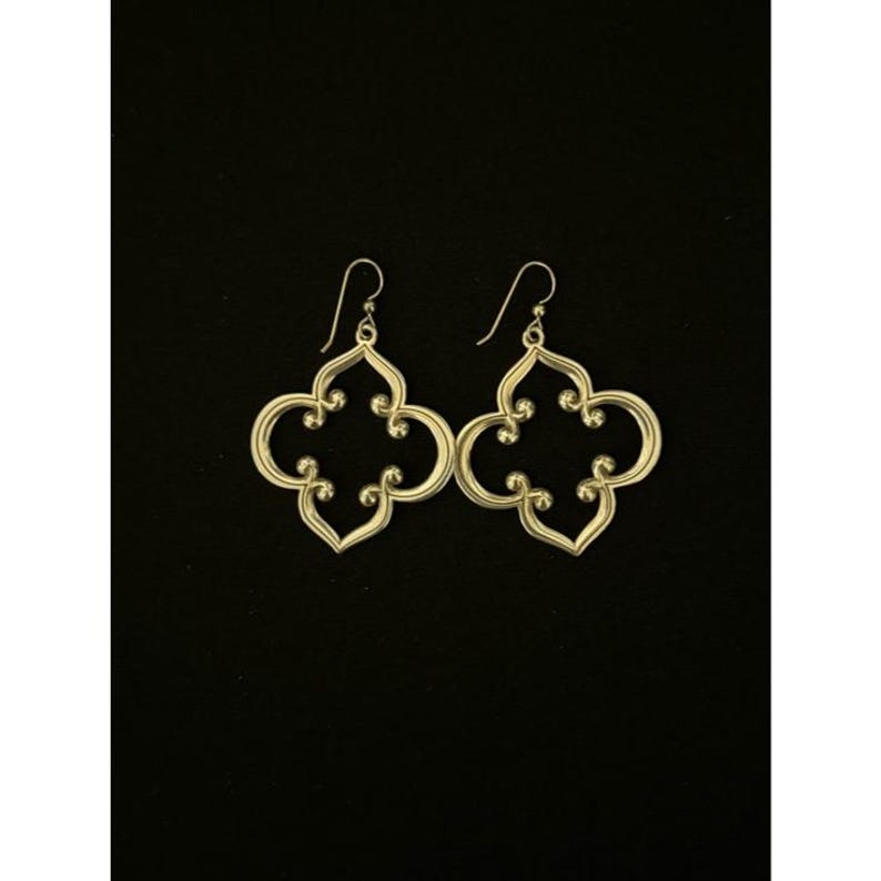 May include: A pair of gold-coloured, openwork earrings with a quatrefoil design. The earrings have a hook closure and are displayed against a black background. The earrings are a decorative accessory.