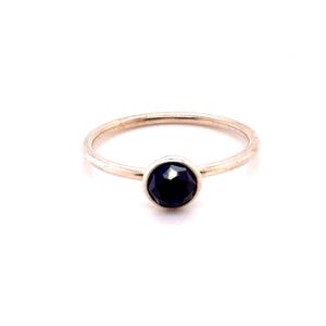May include: A thin, polished silver-tone metal ring featuring a single, round, faceted dark blue gemstone set in a simple bezel setting. The band is smooth and circular, showcasing a minimalist design with a centered, dark blue stone.