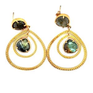 May include: A pair of gold-colored drop earrings with a layered teardrop design. Each earring features a faceted, dark green gemstone at the top and a larger, similar gemstone suspended within the teardrop. The earrings have a textured, dotted border.