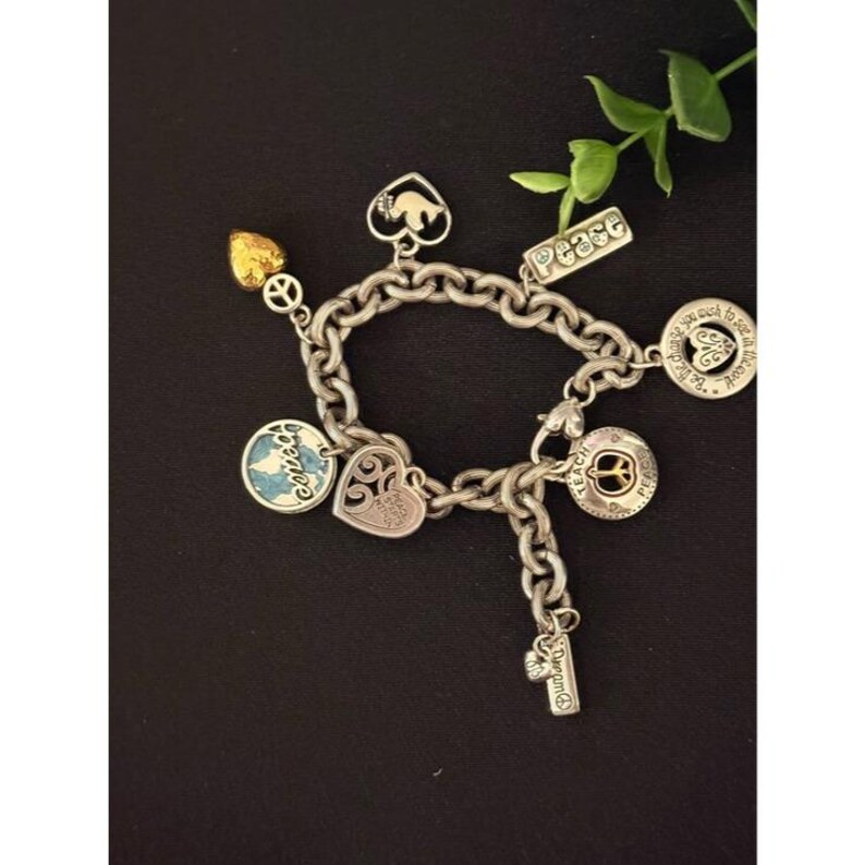 May include: A silver charm bracelet with various charms, including a gold heart, a peace sign, a heart-shaped charm, and a charm with the word "peace". The bracelet is on a black surface.
