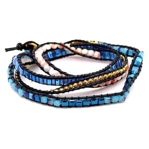 May include: A multi-strand wrap bracelet featuring black leather cord, light blue and dark blue square beads, small metallic gold spheres, and pale pink round beads. The bracelet is coiled in a circular shape with a black leather knot closure.